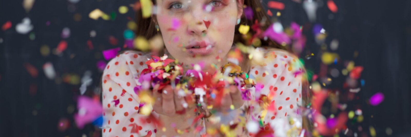 woman blowing confetti in the air isolated over gray